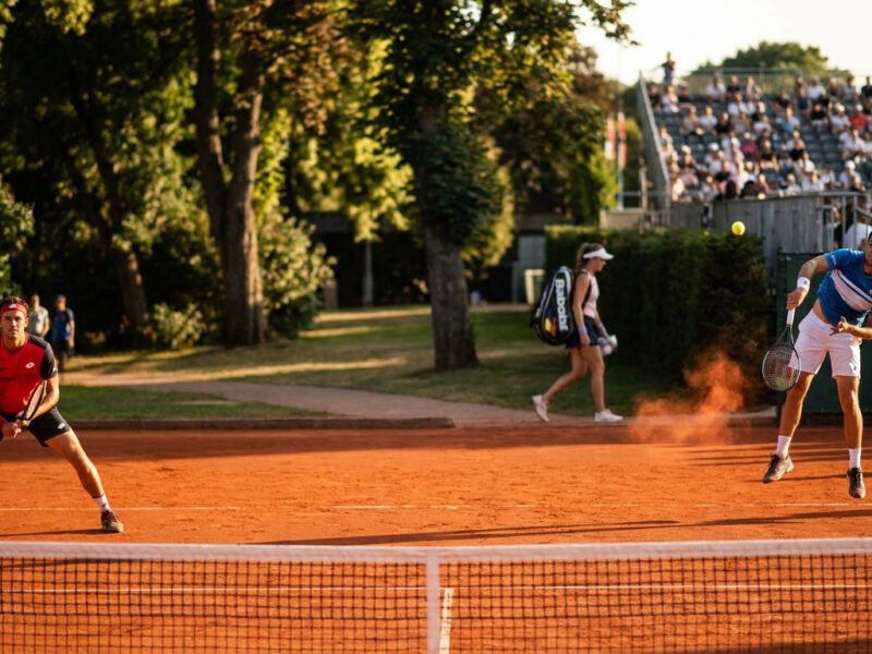 Deux joueurs de tennis en plein échange sur un court en terre battue lors d'un tournoi en extérieur.