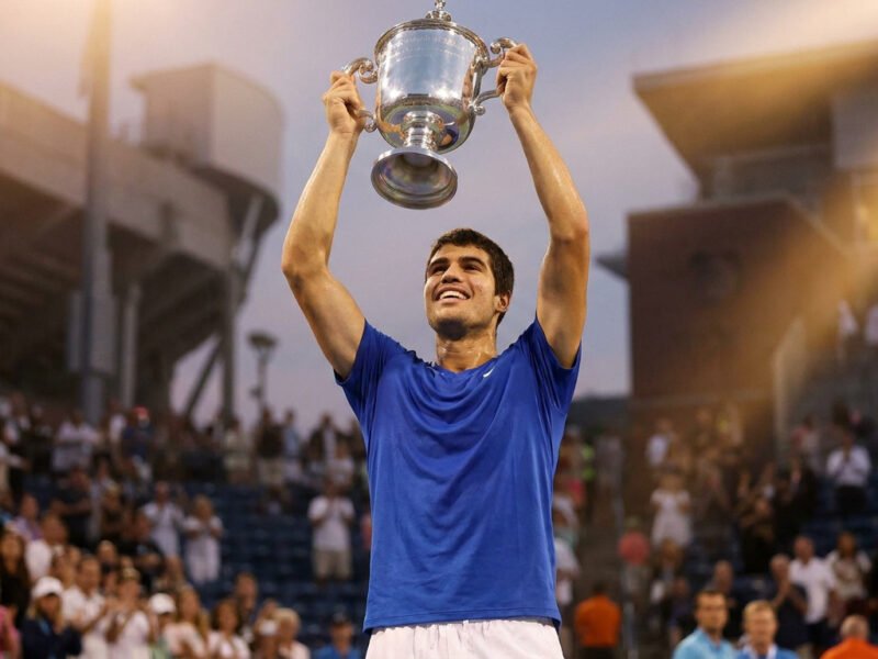 Carlos Alcaraz, en t-shirt bleu, lève fièrement le trophée de l'US Open vers le ciel dans un stade en liesse.